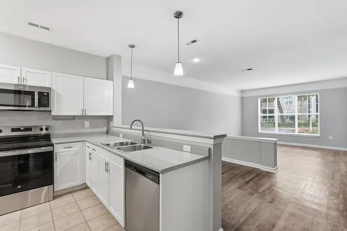 Modern open-concept kitchen and living area with sleek white cabinetry, stainless steel appliances, and elegant pendant lighting, opening to a bright living space with large windows.