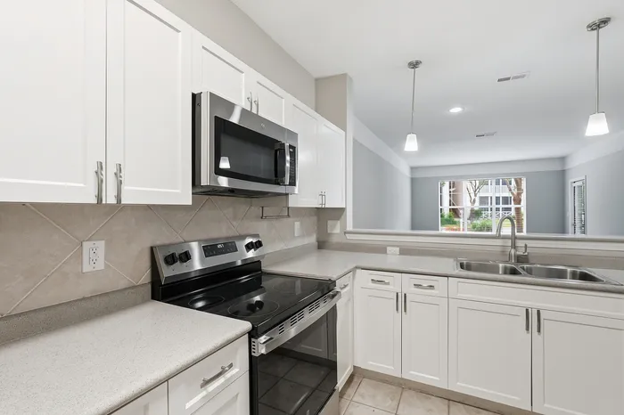 Modern kitchen with sleek white cabinetry, stainless steel appliances, and ample counter space, opening into a bright living area with large windows.