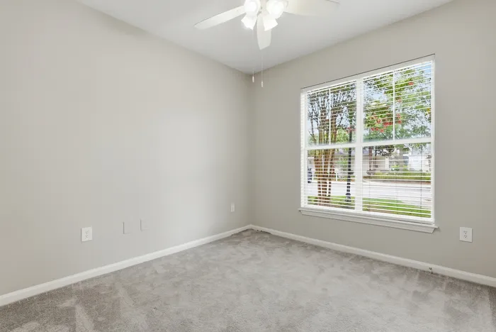 Bright, unfurnished room with plush carpet, large window with blinds, and ceiling fan, perfect for personalizing your living space.
