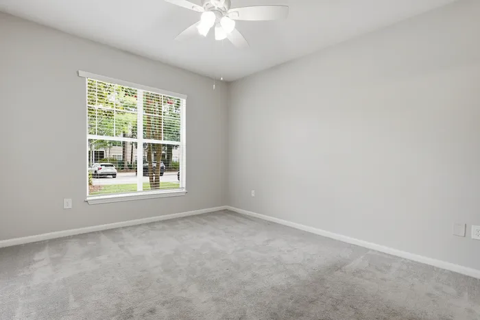 Bright and airy empty room with soft gray carpeting, a large window overlooking greenery, and a modern ceiling fan.