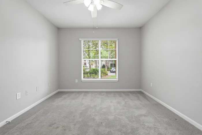 Bright and airy empty room with soft gray carpet, neutral walls, and a large window offering a peaceful view, perfect for a bedroom or office space.