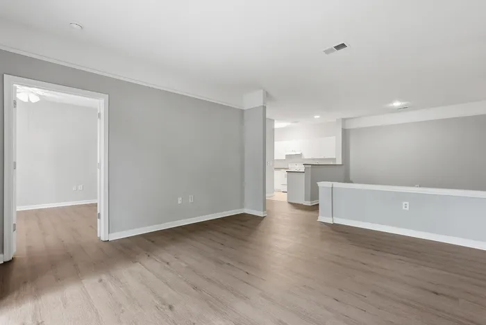 Spacious open-plan living area with sleek wood flooring and neutral-toned walls, leading to a modern kitchen with white cabinetry.