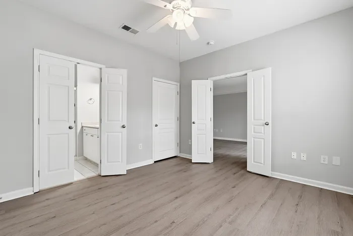 Bright and spacious bedroom with light wood laminate flooring and modern ceiling fan, leading to a pristine white bathroom.