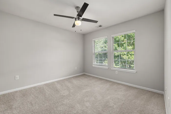 Bright empty room with soft carpet flooring, large windows offering natural light, and a sleek ceiling fan.