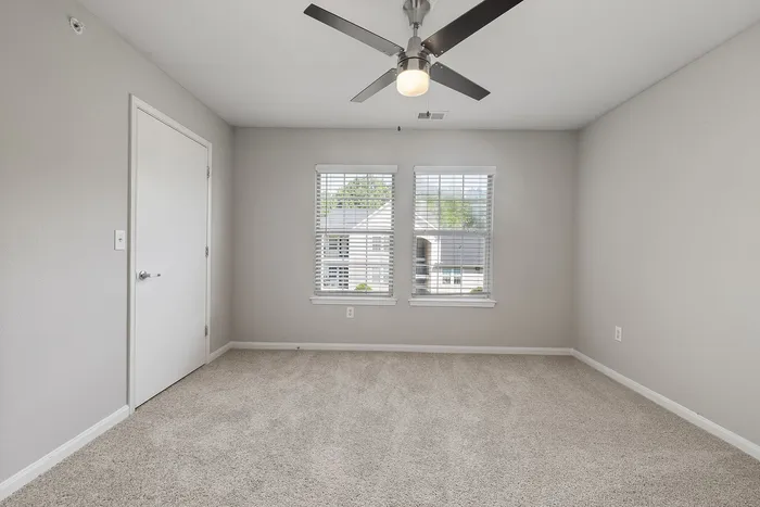 Bright and spacious bedroom with plush carpet, modern ceiling fan, and dual windows offering serene neighborhood views.