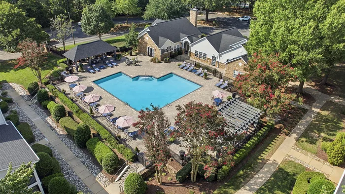 Charming aerial view of a sunny apartment complex featuring a luxurious swimming pool, neat landscaping, shaded lounge areas, and lush greenery.