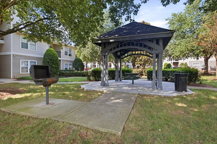 Charming outdoor area with a covered gazebo, barbecue grill, and lush greenery, perfect for social gatherings in a serene apartment community.