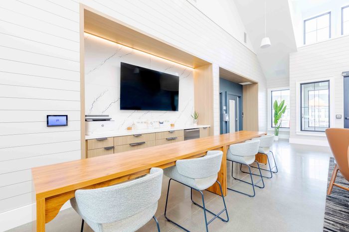 Bright and modern communal space featuring a sleek wooden table, stylish chairs, large wall-mounted TV, and ample natural light from tall windows, creating an inviting environment for gatherings or work.