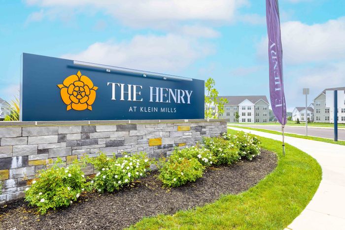 Inviting entrance sign of The Henry at Klein Mills with scenic landscaping and modern apartment buildings in the background.