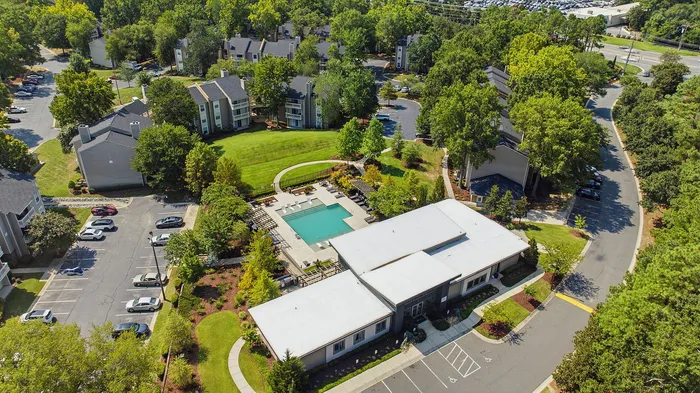 Charming aerial view of a vibrant apartment complex featuring a sparkling pool, lush greenery, and ample parking surrounded by serene natural beauty.