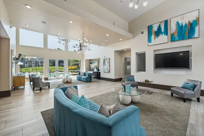 Elegant living room with high ceilings, modern chandeliers, and large windows overlooking scenic greenery. Features stylish teal and gray furniture for a luxurious ambiance.