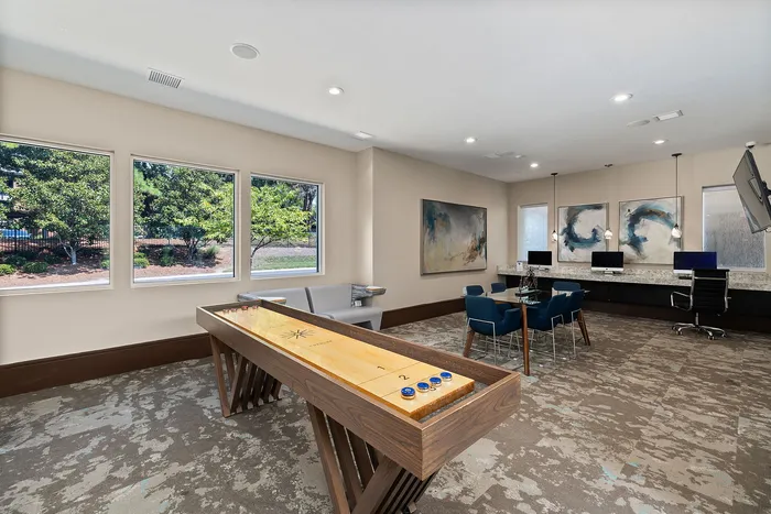 Modern apartment community room with large windows, shuffleboard table, and cozy seating areas, perfect for social gatherings and leisure.