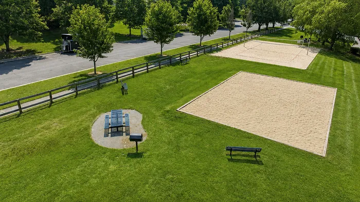 Inviting outdoor recreation area with two sand volleyball courts, picnic table, and grilling spot surrounded by lush green lawns and trees.