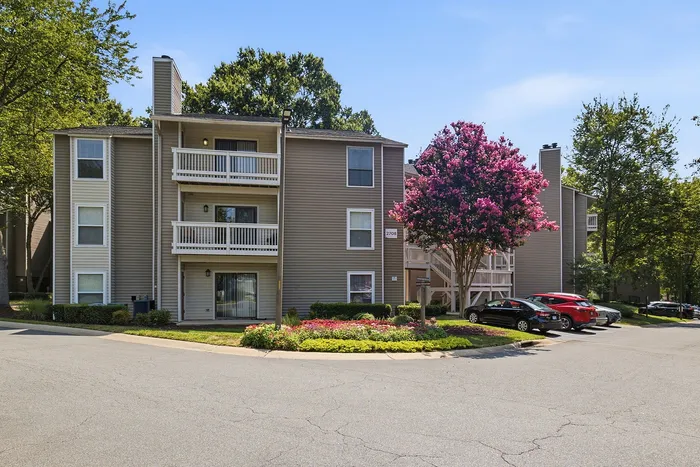 Charming apartment complex with three levels, private balconies, lush landscaping, and vibrant pink trees under a clear blue sky.