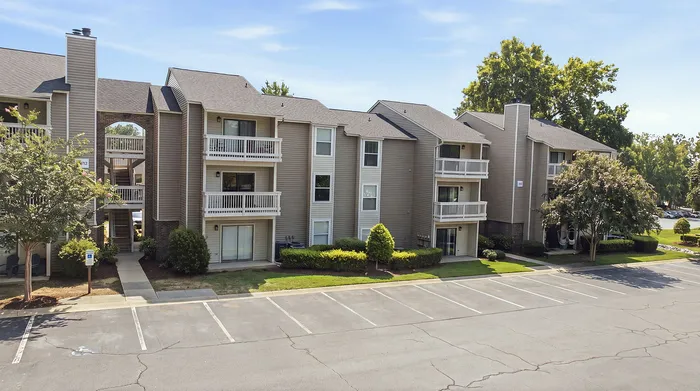 Charming three-story apartment building with private balconies, manicured landscaping, and ample parking space under a clear blue sky.