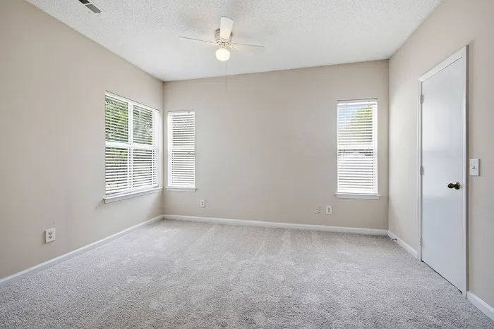 Bright and airy bedroom with plush carpeting, ample natural light from large windows, and a sleek ceiling fan.