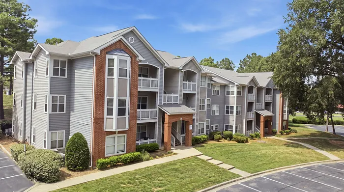 Charming apartment complex with modern gray and brick facade, surrounded by lush greenery and ample parking space, perfect for comfortable living.