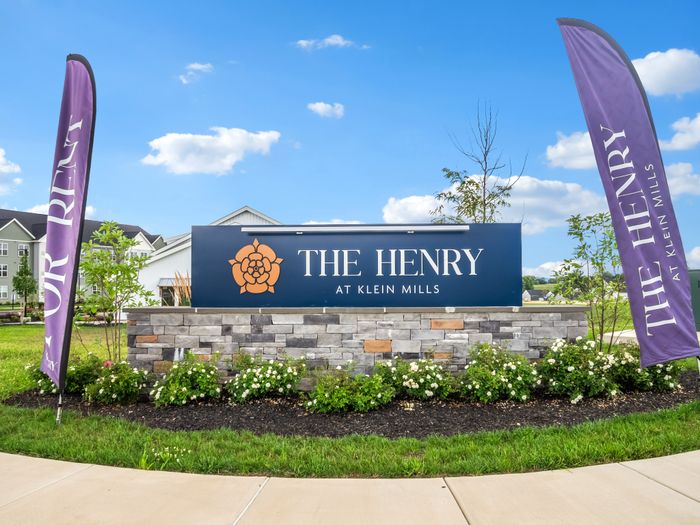 Inviting sign of The Henry at Klein Mills with beautiful landscaping and vibrant flags under a clear blue sky.