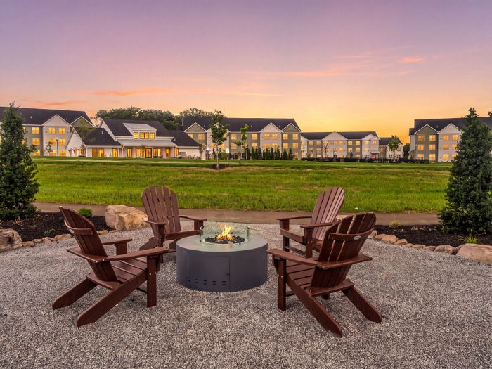 Charming outdoor area with cozy fire pit surrounded by wooden chairs, overlooking elegant apartments under a stunning sunset sky.