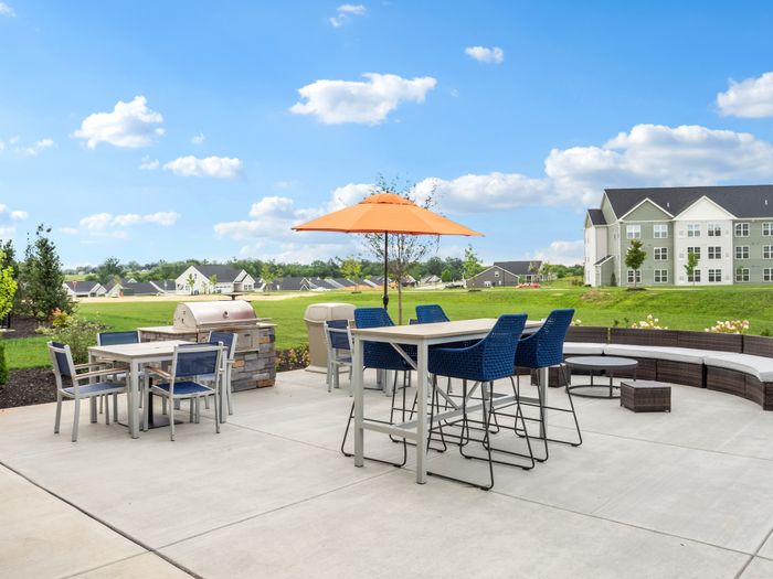 Charming outdoor patio with stylish seating, modern grill, and vibrant orange umbrella against a scenic backdrop of lush green lawns and elegant apartment buildings.