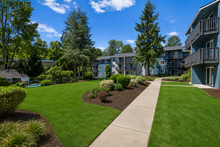 Charming apartment exterior with manicured lawns, vibrant gardens, and a tranquil pool area under a bright blue sky.