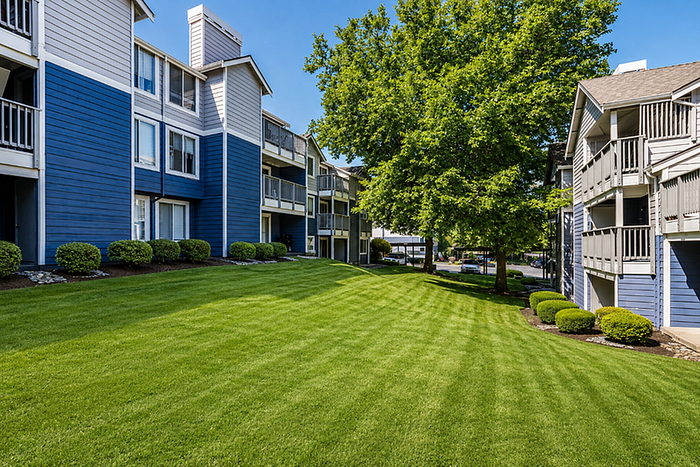 Charming apartment complex featuring modern blue buildings with balconies, lush green lawn, and serene trees.