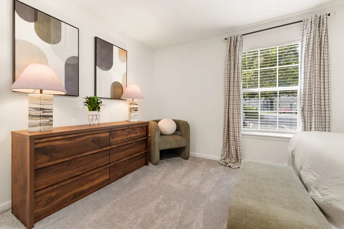 Elegant bedroom with cozy modern decor, featuring a stylish wooden dresser, ambient lighting, and large window with serene outdoor views.