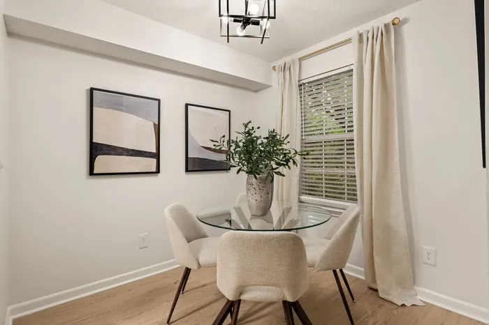 Elegant dining area with a modern glass table, cozy beige chairs, artistic wall decor, and a large window with flowing curtains.