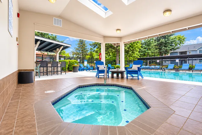Inviting indoor-outdoor pool area featuring a sparkling hot tub, comfortable lounge seating, and vibrant greenery under a bright, skylit ceiling.