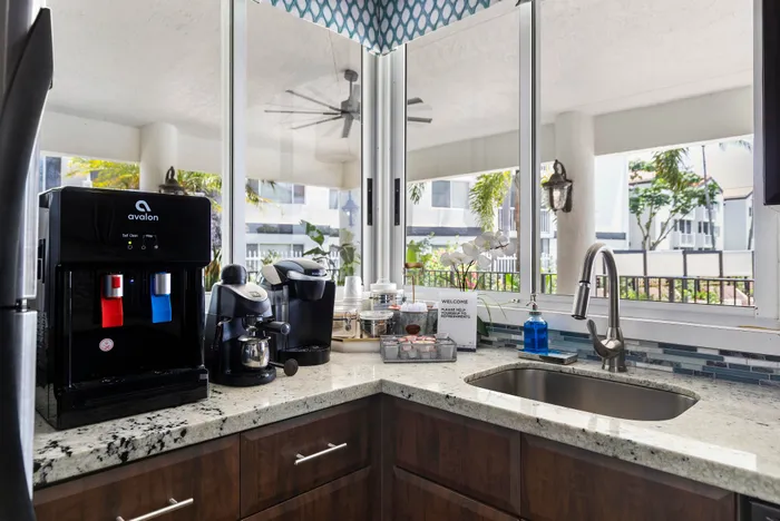 Modern kitchen corner with granite countertops, luxury coffee machines, and a stainless steel sink. Bright windows offer a lush outdoor view, creating an inviting atmosphere.