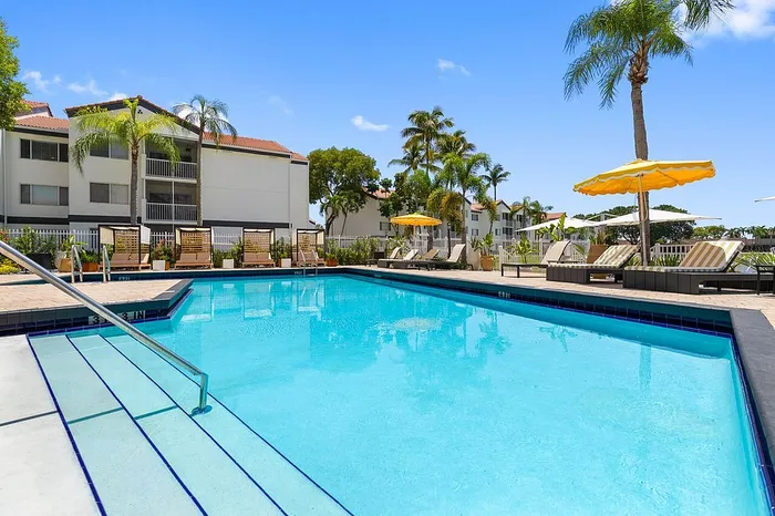 Inviting outdoor pool area at a modern apartment complex, featuring palm trees, sun loungers, and bright yellow umbrellas beneath a clear blue sky.