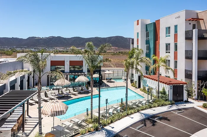Luxurious apartment complex with a sparkling pool, sun loungers, and palm trees, set against a scenic mountainous backdrop.