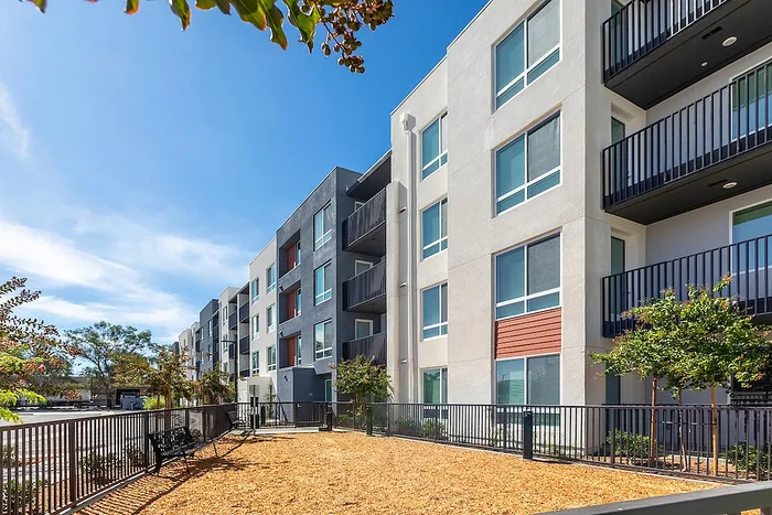 Modern apartment complex with sleek design, spacious balconies, and lush landscaping under a bright blue sky.