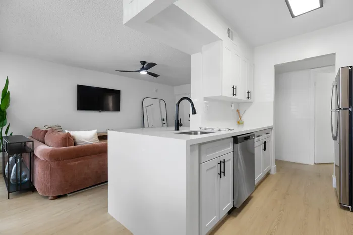 Contemporary apartment kitchen with sleek white cabinets, modern stainless steel appliances, and an inviting open layout leading to a cozy living room with a plush sofa and wall-mounted TV.