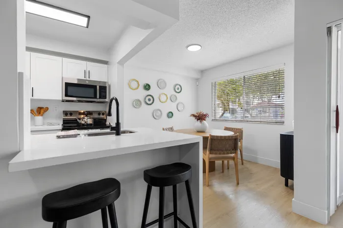 Modern kitchen and dining area featuring sleek white cabinetry, stylish breakfast bar with stools, and a cozy dining nook with window views.