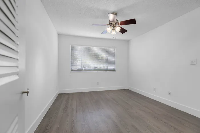 Bright and airy room with elegant hardwood floors, large window with blinds, and a modern ceiling fan.