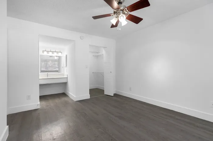 Modern bedroom with sleek hardwood floors, a ceiling fan, and a spacious open closet, featuring a bright vanity area with a large mirror for a welcoming touch.