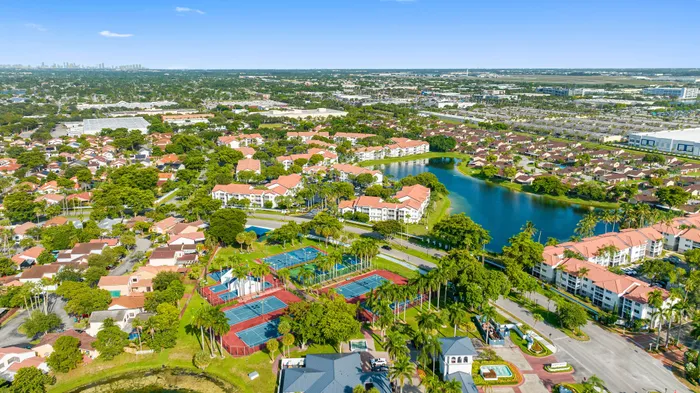 Aerial view of lush residential community featuring charming homes with red-tiled roofs, surrounded by serene lakes and vibrant greenery, complete with tennis courts and a scenic landscape.