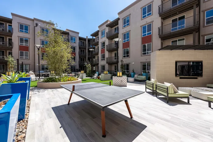 Modern apartment courtyard featuring elegant seating areas, decorative planters, and a stylish ping pong table under a clear blue sky.