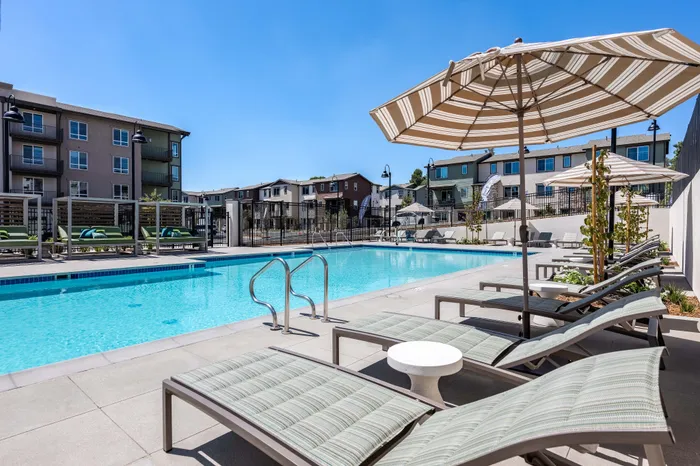 Luxurious apartment pool area with modern lounge chairs, large umbrellas, and a scenic view of contemporary buildings.