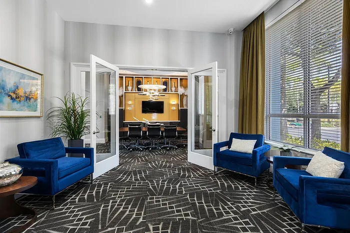 Elegant apartment interior with French doors leading to a chic conference room, featuring plush blue armchairs, vibrant artwork, and abundant natural light.