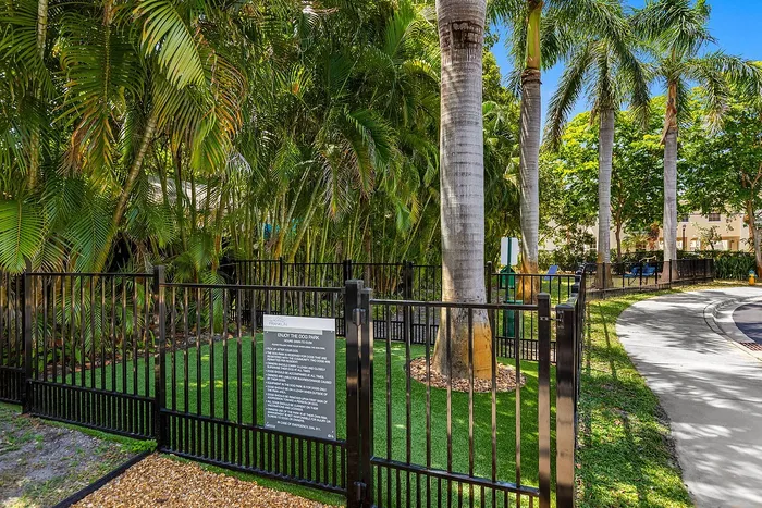 Lush tropical dog park enclosed by a stylish black fence, surrounded by tall palm trees and a paved walkway, offering a serene outdoor retreat.