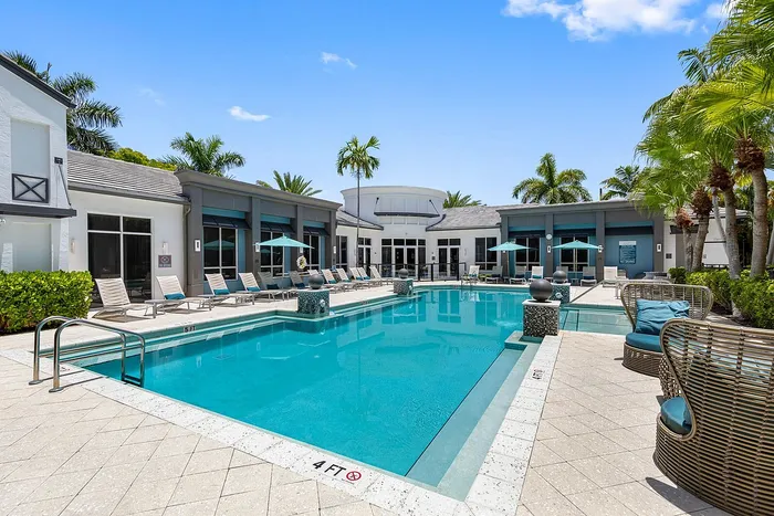 Luxurious apartment community pool area with sparkling water, surrounded by stylish lounge chairs and palm trees under a clear blue sky.