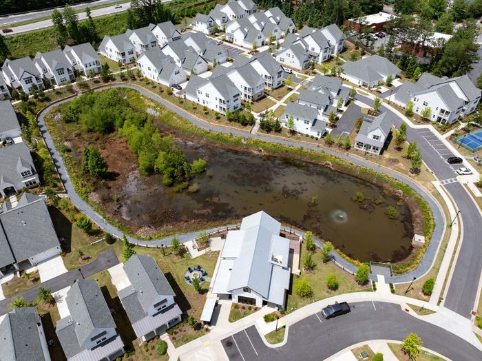 Aerial view of a picturesque residential community featuring charming houses with grey roofs, a central lush green pond, and inviting walking paths.