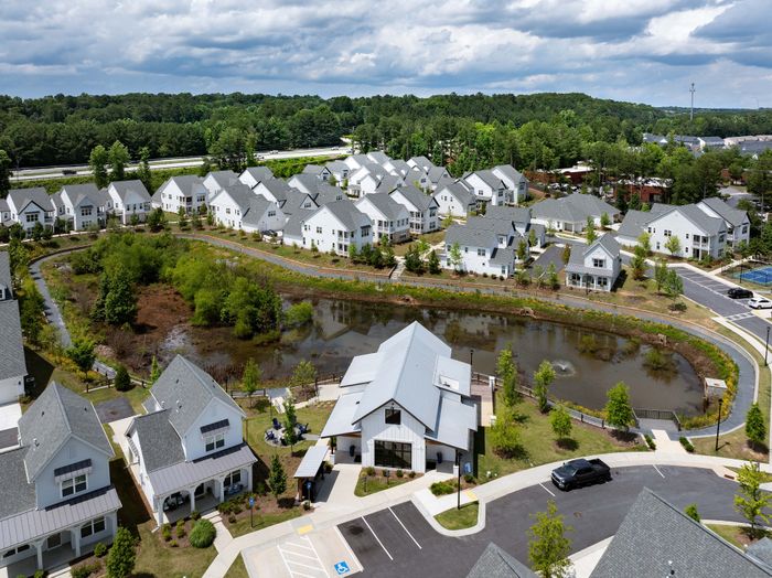 Charming aerial view of a serene residential community with modern white houses, surrounding a picturesque pond and lush greenery, promising a peaceful lifestyle.