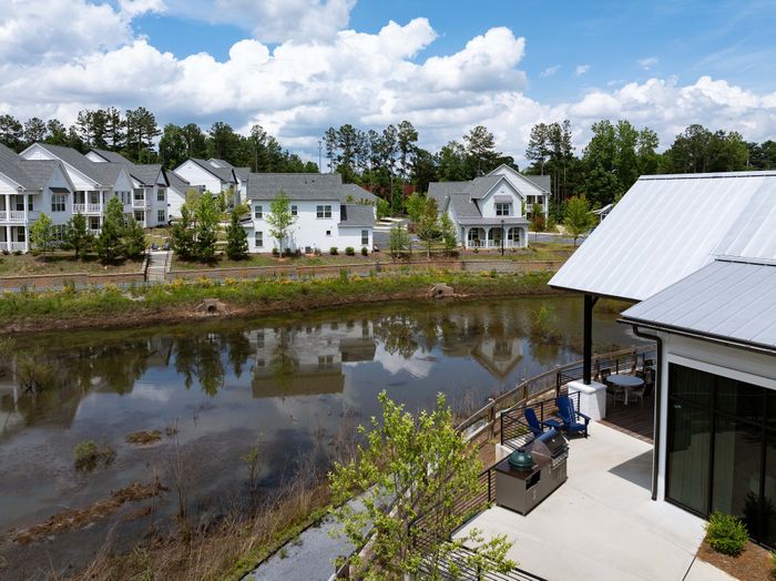 Charming lakeside apartment view with stylish patio, nestled among elegant homes and lush greenery under a vibrant blue sky.