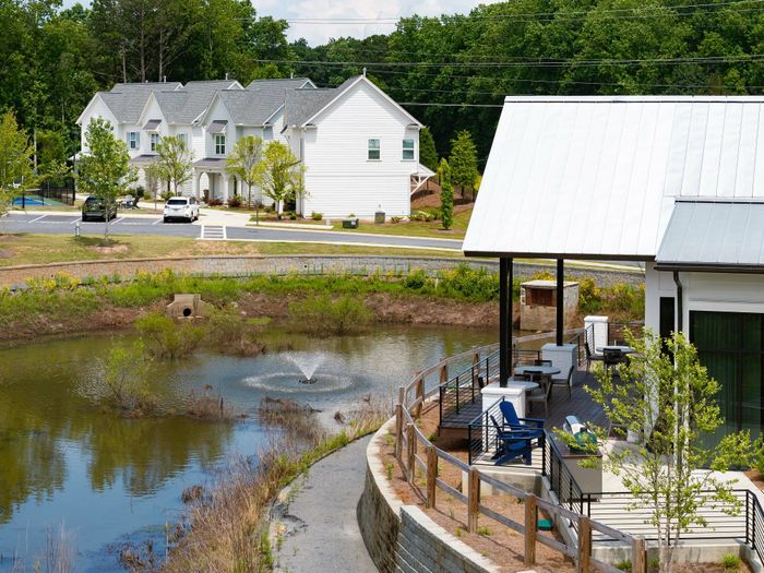 Charming suburban scene showcasing elegant townhouses with lush greenery, a tranquil pond, and a cozy patio area under a modern roof.