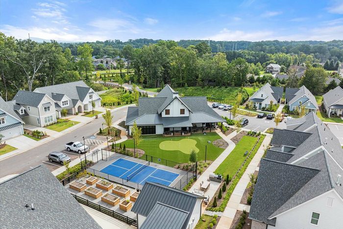Charming suburban neighborhood with modern homes, lush greenery, a community center, putting green, and tennis court under a bright blue sky.