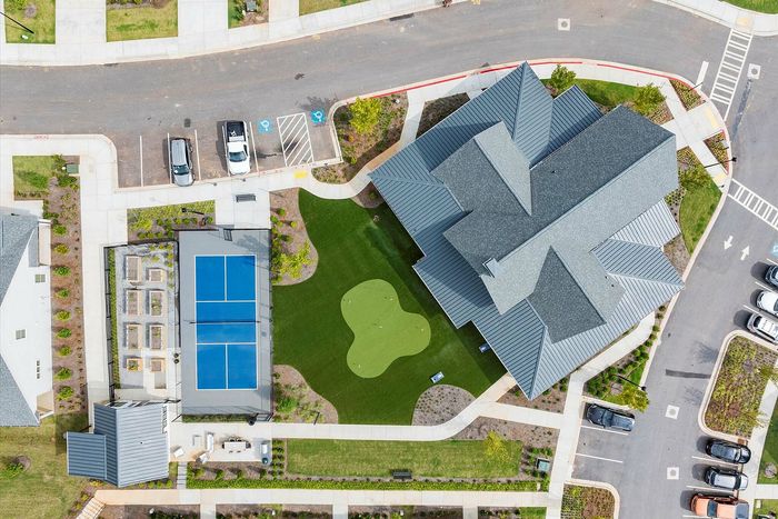 Aerial view of a modern housing complex featuring a vibrant blue tennis court, lush green lawns, and ample parking surrounded by well-maintained landscaping.