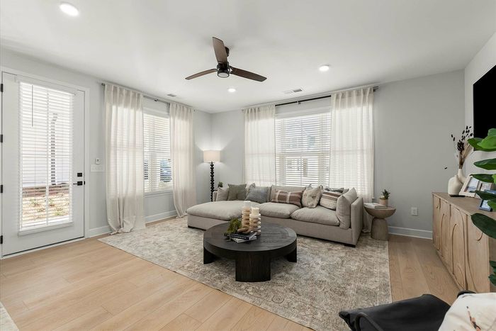Bright and inviting living room with a modern beige sofa, elegant floor lamp, and natural light streaming through large windows with sheer curtains. Stylish round coffee table and light wood flooring create a cozy and chic ambiance.